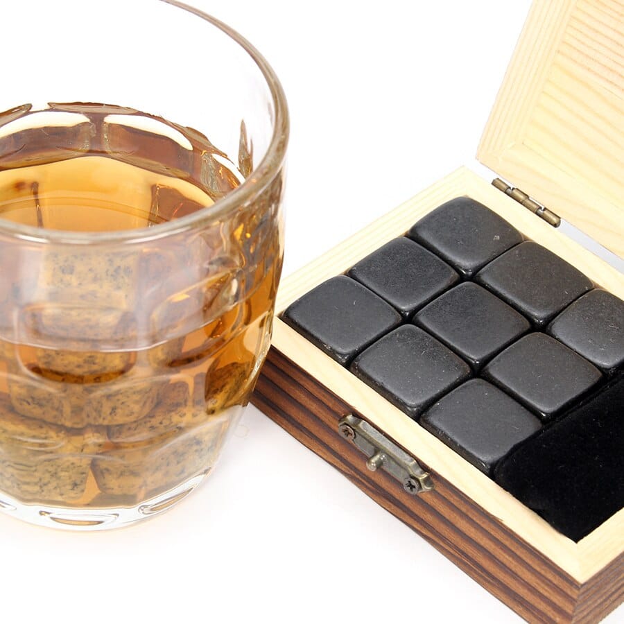 Coffret pierre à whisky en bois ouvert avec neuf pierres noires à côté d'un verre de whisky ambré