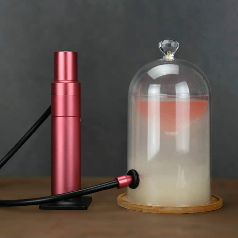 Smoking gun rouge remplissant de fumée une cloche en verre posée sur un plateau bois près d'un cocktail