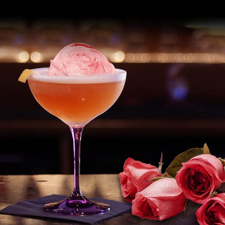 Glaçon en forme de rose dans une coupe à cocktail orangée, roses roses en décor sur un bar tamisé