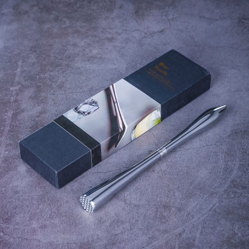 Pilon cocktail design en acier chromé argenté posé près de son coffret cadeau noir sur un plan en béton gris