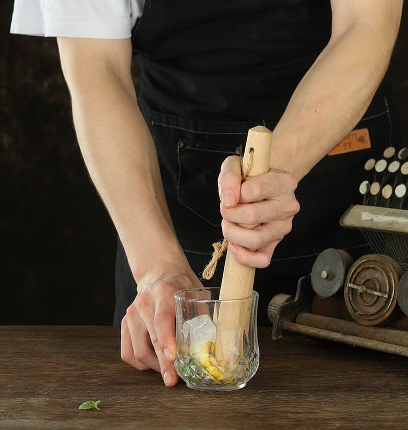 Barman écrasant citron et menthe dans un verre à facettes avec le pilon en bois clair à cordelette en jute