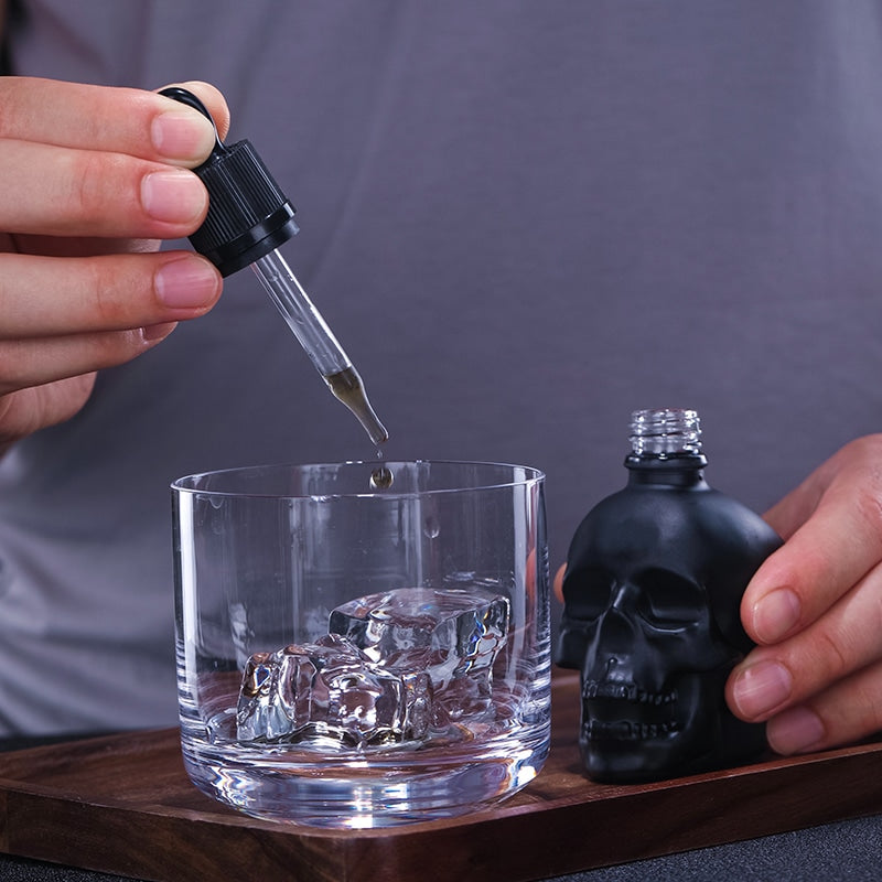 Pipette à cocktail tête de mort noire déposant une goutte dans un verre à glaçons sur un plateau bois