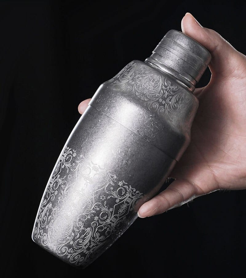 Shaker Cocktail Professionnel cobbler en acier argent vieilli à arabesques gravées, tenu en main sur fond noir