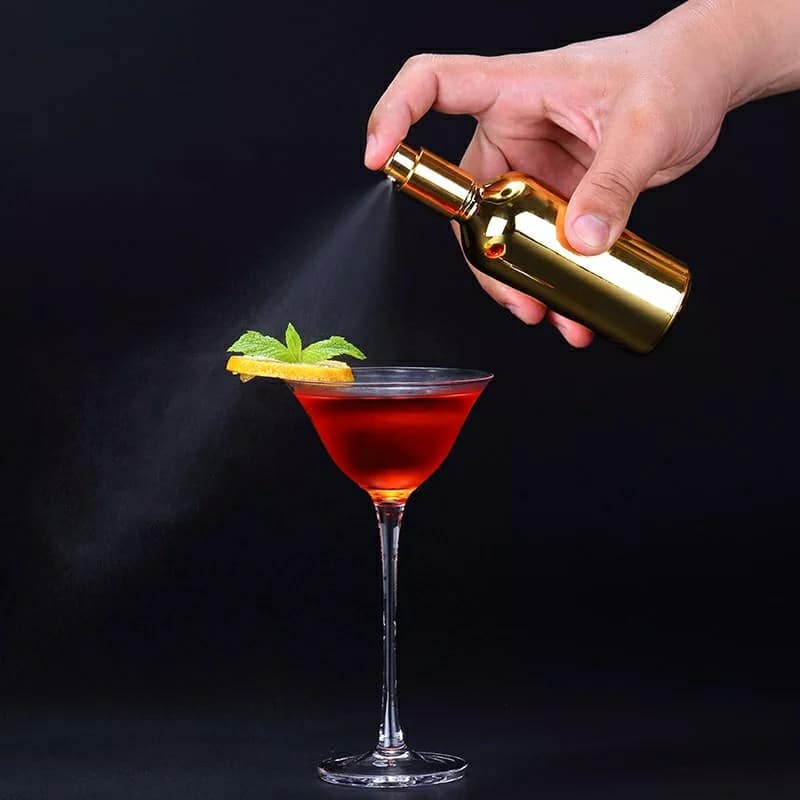 Spray pulvérisateur culinaire design doré vaporisant une brume sur un cocktail rouge en verre à martini