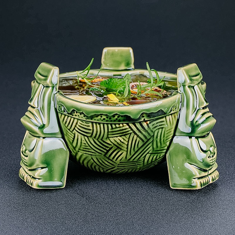 Récipient en céramique tiki vert porté par trois statues moaï, rempli d’un cocktail garni de menthe et de romarin