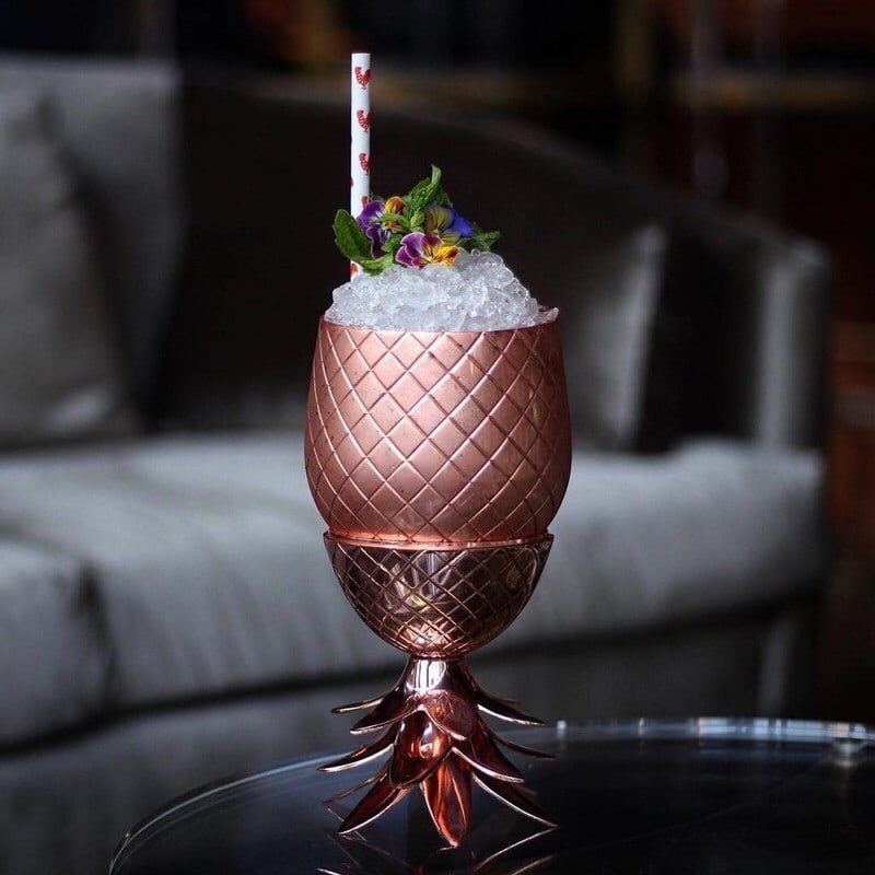 Verre Moscow Mule Ananas cuivré sur pied à feuilles, glace pilée, paille et fleurs comestibles en garniture