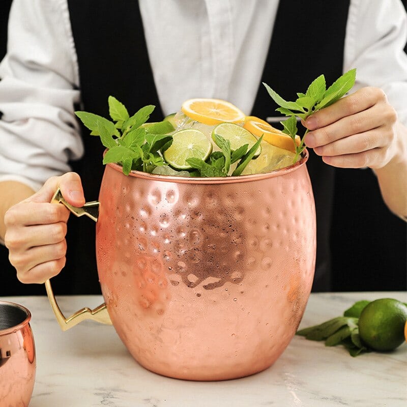 Verre Moscow Mule Géant en cuivre martelé à anse dorée, tenu par un barman et garni de citrons, menthe et glace