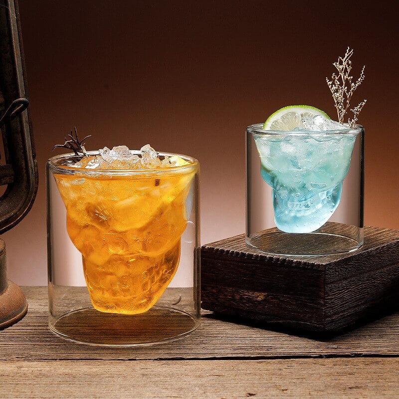 Deux verres whisky tête de mort double paroi, cocktail orange et cocktail bleu au citron vert, posés près d’une lanterne ancienne