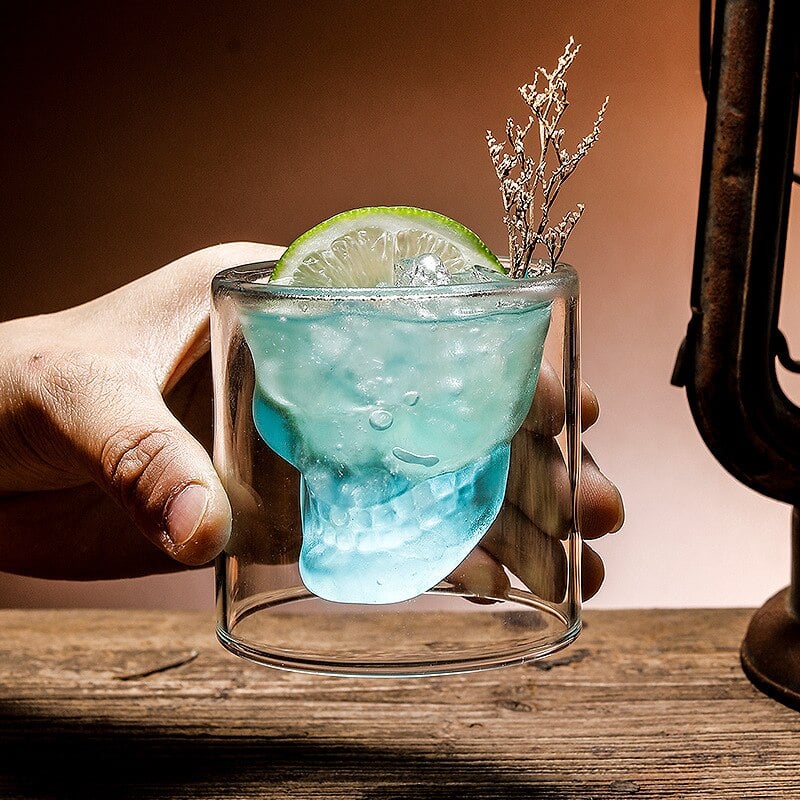 Main tenant un verre whisky tête de mort avec cocktail bleu, rondelle de citron vert et fleurs séchées