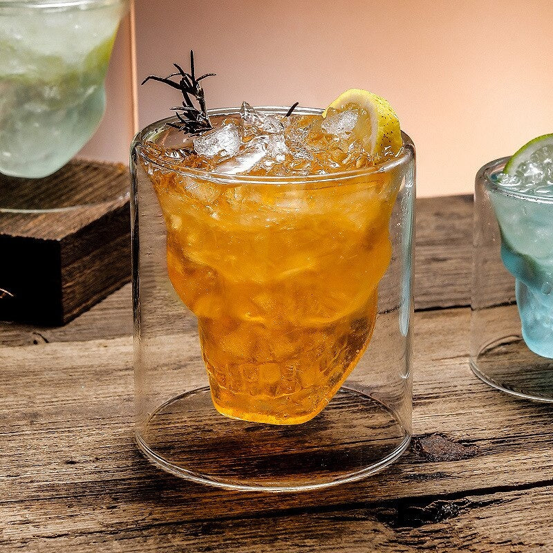 Verre à whisky tête de mort rempli d’un cocktail ambré avec glace pilée, rondelle de citron et brin de romarin, sur bois brut