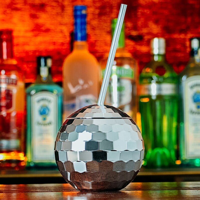 Verre à Cocktail Disco boule à facettes miroir argentée avec couvercle et paille, posé au bar parmi les bouteilles