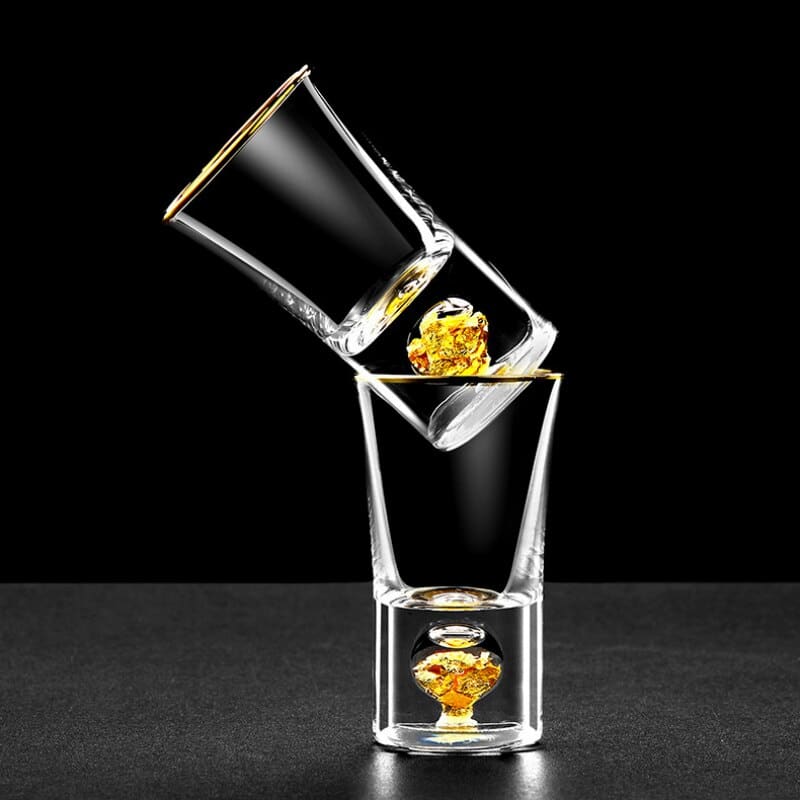 Verres à Shot Or à rebord doré empilés en biais, feuilles d'or visibles dans le fond, fond noir