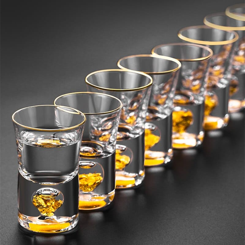 Verres à Shot Or à rebord doré alignés, feuilles d'or dans le fond épais, alcool clair sur fond noir