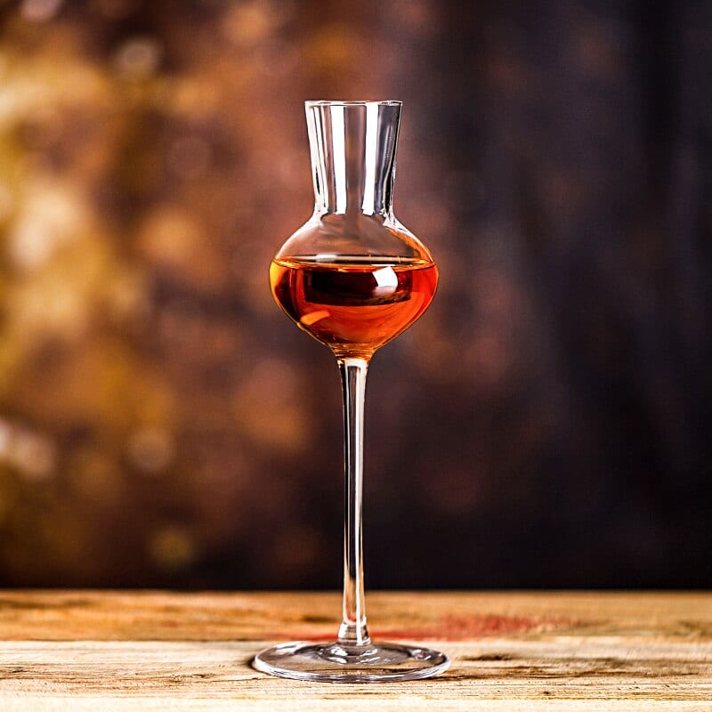 Verre à whisky long à pied fin, calice bulbeux rempli d’un spiritueux ambré, posé sur une table en bois