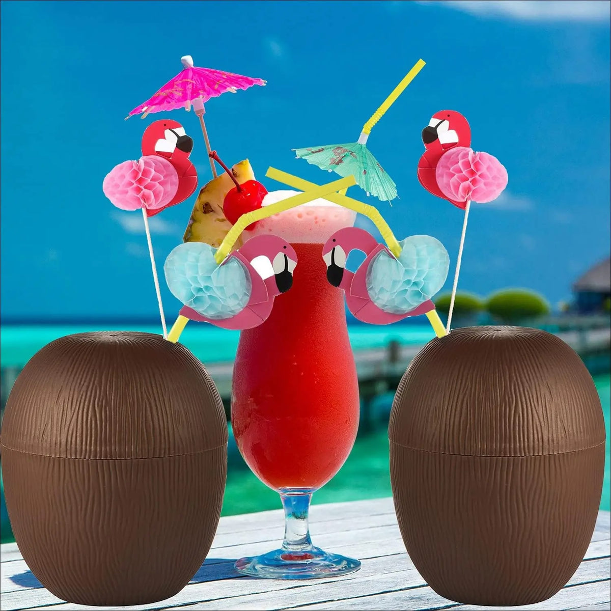 Deux verres noix de coco encadrant un cocktail rouge sur un ponton, lagon turquoise en arrière plan