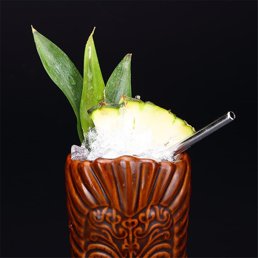 Verre Tikinator brun rempli de glace pilée, tranche d'ananas, feuilles et paille métal, fond noir