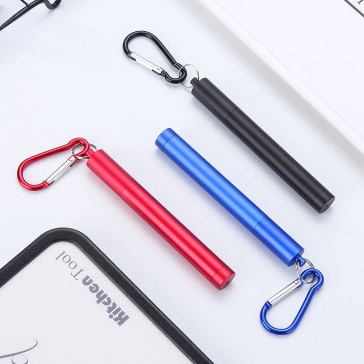 Trois étuis de paille inox rétractable à mousqueton en noir, rouge et bleu posés sur fond clair