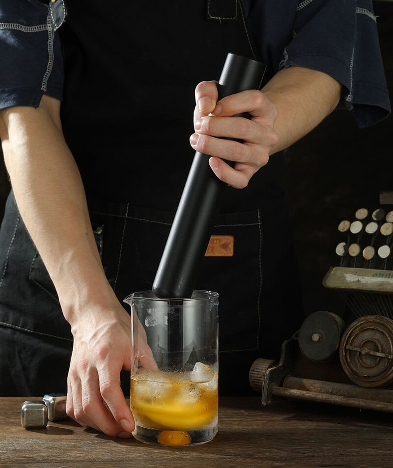 Barman écrasant orange, citron et glace dans un verre avec le pilon pour cocktail noir mat cylindrique