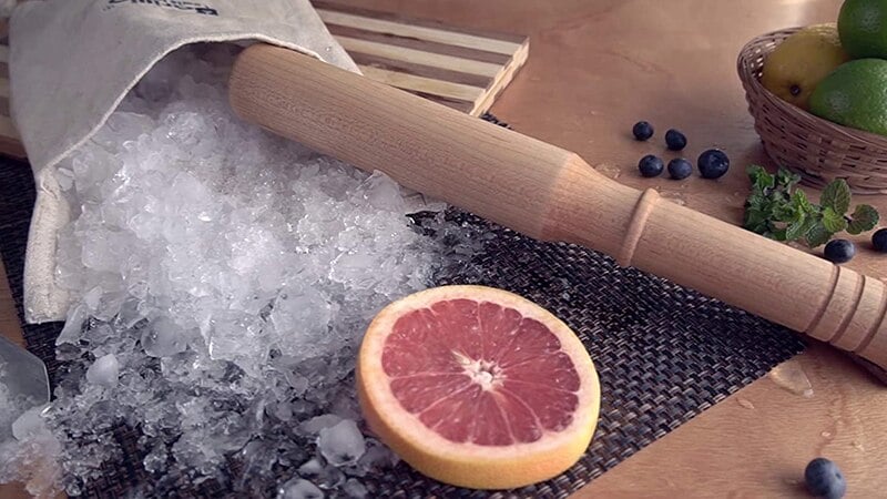 Pilon cocktail en bois en scène avec glace pilée, demi pamplemousse, myrtilles, citrons verts et menthe