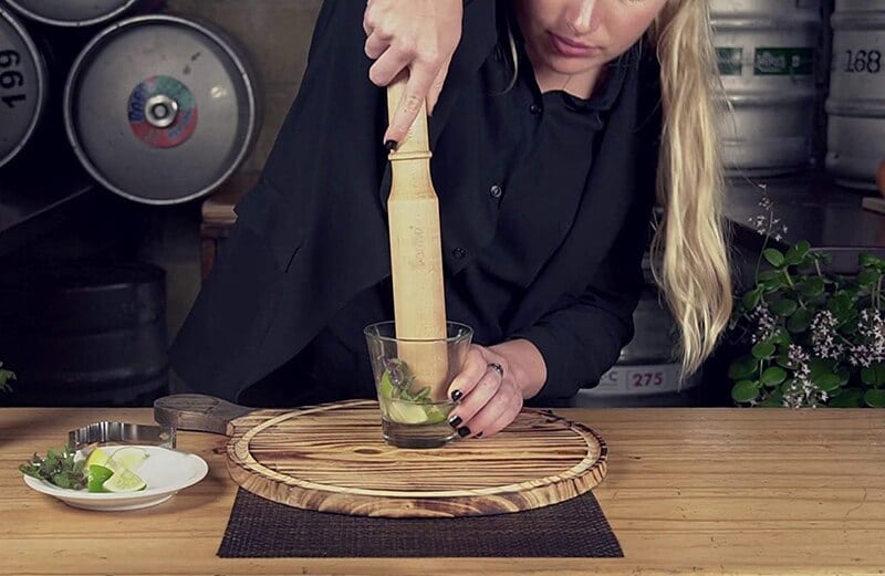 Femme écrasant menthe et citron vert dans un verre avec le pilon cocktail en bois en ambiance de bar