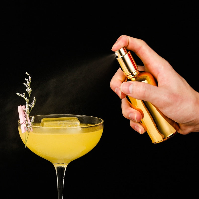 Spray pulvérisateur culinaire design doré vaporisant une brume sur un cocktail jaune fleuri en coupe