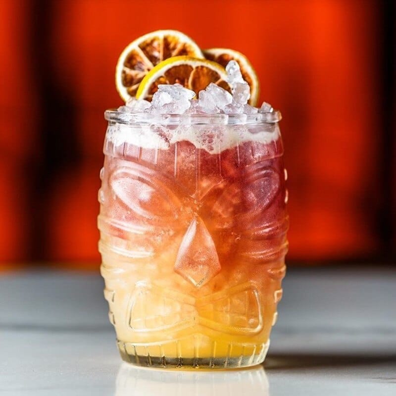Tiki Verre transparent rempli d'un cocktail rouge et ambré, glace pilée et agrumes séchés, fond rouge