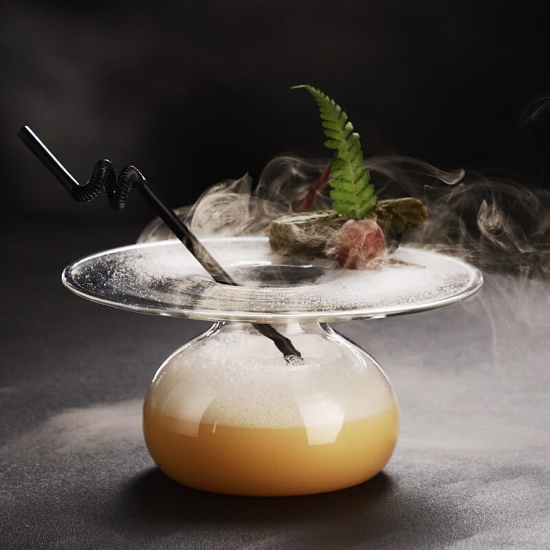 Verre à Cocktail Géant XXL soucoupe avec cocktail ambré mousseux, fumée de glace carbonique et fougère, fond noir