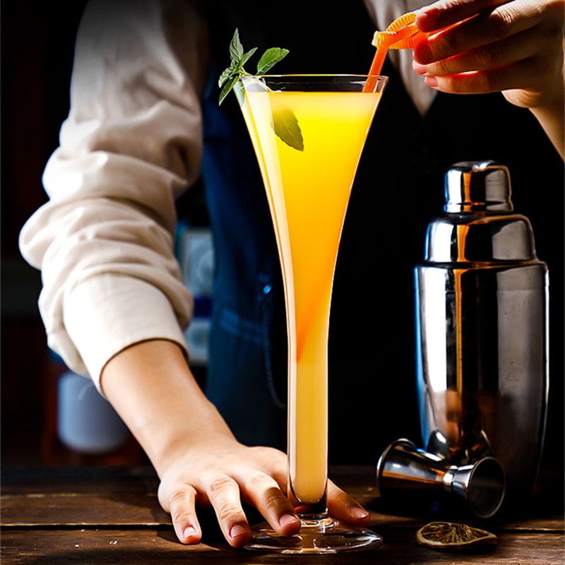 Verre cocktail trompette rempli d’un cocktail orange à la menthe, posé par un barman à côté d’un shaker