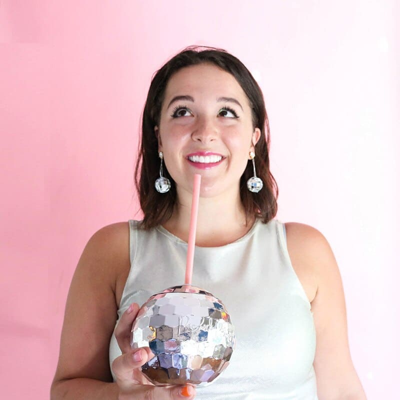 Verre à Cocktail Disco boule miroir tenu par une femme souriante avec paille rose sur fond rose