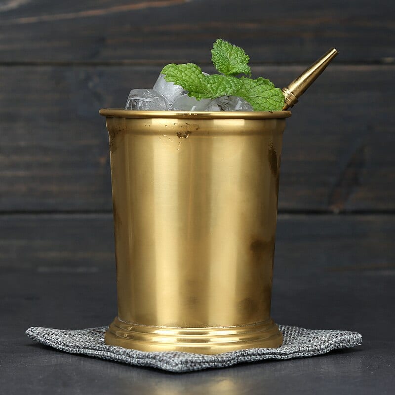 Verre Moscow Mule Julep doré laiton rempli de glace et de menthe, paille dorée, posé sur un dessous gris