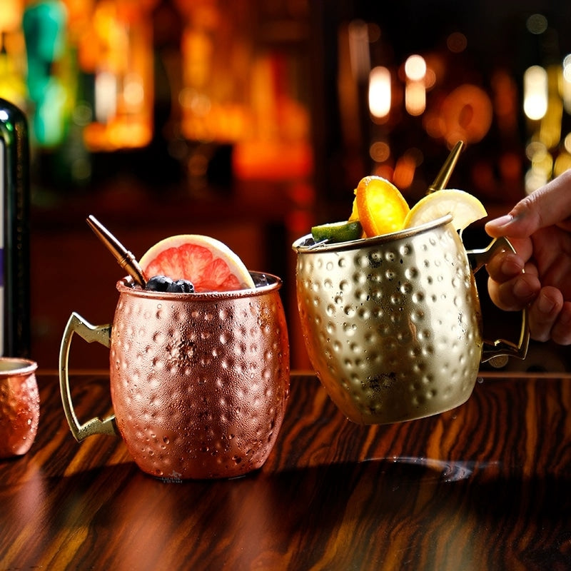 Main saisissant un verre Moscow Mule or garni d’agrumes, à côté d’un mug cuivré, dans un bar tamisé