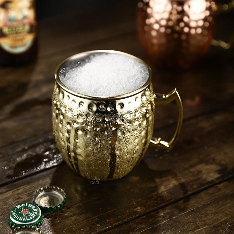 Verre Moscow Mule or en inox martelé rempli d’une boisson mousseuse, posé sur une table en bois avec des capsules