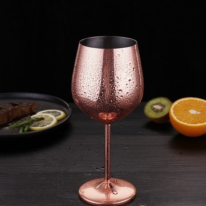 Verre à vin original rose gold couvert de gouttes, dressé près d’une assiette, d’un kiwi et d’une orange