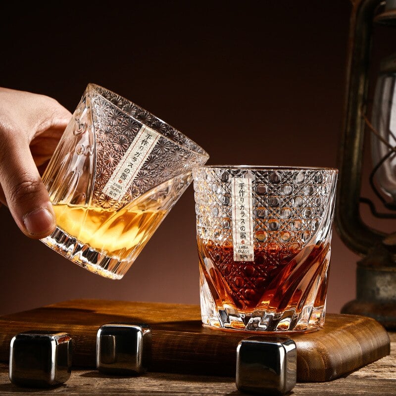 Deux verres whisky luxe ciselés qui trinquent au dessus de pierres à whisky en inox, ambiance de bar tamisée