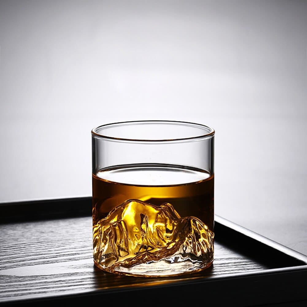 Verre whisky montagne rempli de whisky ambré, relief de montagne sculpté au fond, posé sur un plateau en bois noir