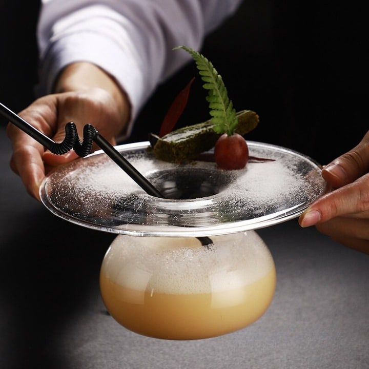 Verre à Cocktail Géant XXL soucoupe transparent, cocktail ambré mousseux et fougère, présenté à deux mains