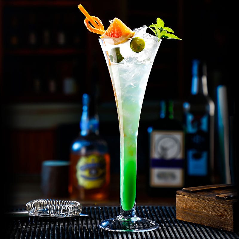 Verre cocktail trompette garni d’un cocktail vert glacé avec pamplemousse, olives et menthe dans un bar tamisé