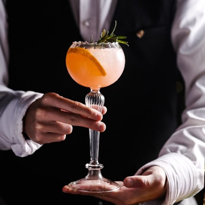Verre à Cocktail Vintage à pied cannelé tenu par un barman, cocktail pamplemousse et romarin, gilet noir