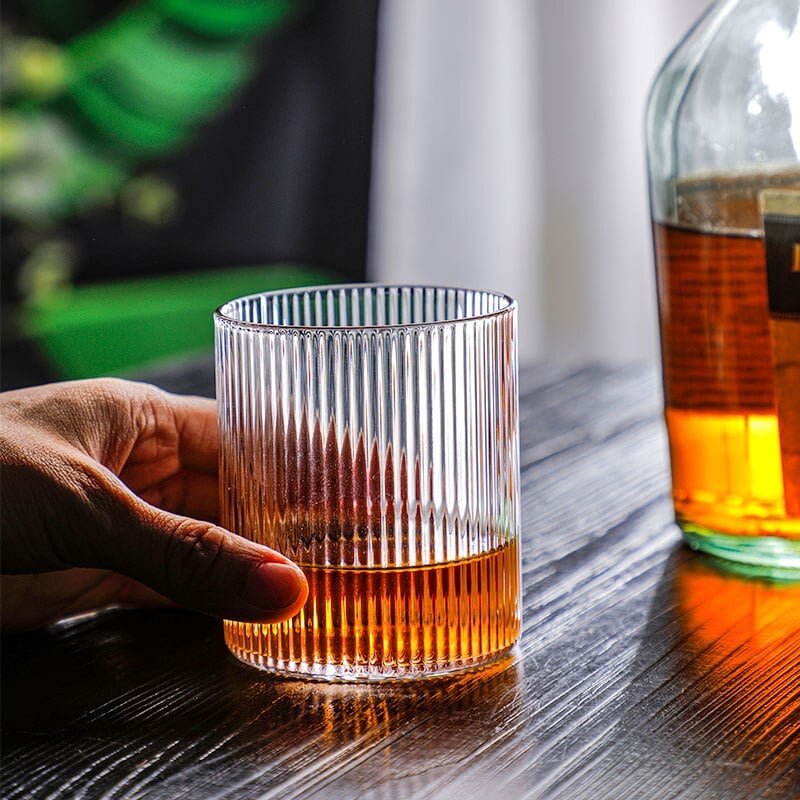 Main posée sur un verre à whisky design strié rempli de whisky, bouteille de whisky en arrière plan