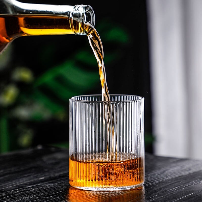 Whisky versé d’une bouteille dans un verre à whisky design aux stries verticales, sur une table en bois noir