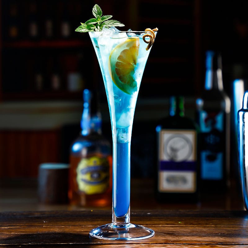 Verre cocktail trompette contenant un cocktail bleu avec rondelle de citron et menthe, sur un comptoir en bois